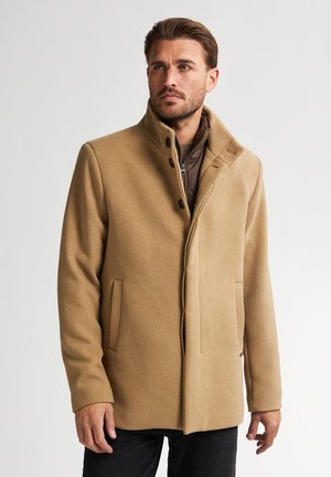 Beige wollen jas met hoge kraag, enkele knoopsluiting, zwarte knopen en zijzakken. Gedragen over een bruine binnenlaag.
