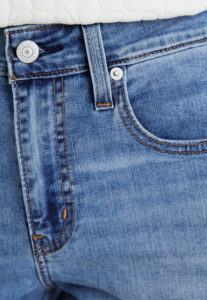 Hellblaue Jeans aus Denim mit einem ausgebleichten Look, ausgestattet mit einem Knopfverschluss, orangen Nähten und einer Vordertasche auf der rechten Seite.
