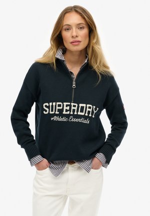 Pull à zip noir marine avec le texte blanc "SUPERDAY Athletic Essentials", associé à une chemise à col rayée en dessous. Tissu tricoté texturé.