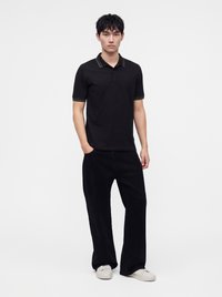 Jeune homme debout, les mains dans les poches, portant un polo noir avec un col rayé et un pantalon noir avec des baskets blanches.