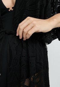Kimono in pizzo nero con intricate fantasie, apertura frontale e tessuto leggero. Chiusura a cravatta regolabile in vita.