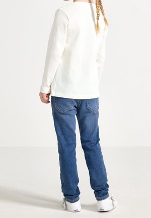 Weißes, langärmliges, geripptes Shirt kombiniert mit blauem Denim-Jeans und weißen Turnschuhen. Das Outfit zeichnet sich durch ein schlichtes Design ohne sichtbare Muster aus.