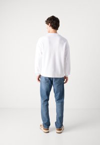 Camisa blanca de manga larga con puños acanalados, combinada con jeans azul claros y sandalias marrones, vista desde atrás contra un fondo liso.