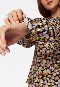 Frau, die den Bund einer blumenmusternden Bluse in lila, orange, weiß und schwarz auf weißem Hintergrund anpasst.