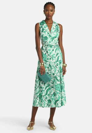 Mujer con vestido midi sin mangas con cuello y estampado floral verde, sosteniendo un bolso de mano verde texturizado, con pulseras doradas y tacones dorados.