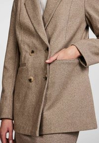 Kobieta ubrana w beżową dwurzędową marynarkę z czterema guzikami, z jedną ręką w kieszeni, dopasowaną spódnicę lub spodnie oraz białą bluzkę pod spód.