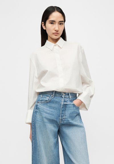 Λευκός πουκάμισος με μακριά μανίκια και κουμπιά, με γιακά, σε συνδυασμό με ανοιχτό μπλε τζιν παντελόνι φαρδιάς γραμμής. Το υλικό φαίνεται λείο, άνετη εφαρμογή, ελάχιστες λεπτομέρειες.
