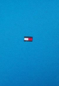 Tissu turquoise au look texturé avec un petit logo brodé en rayures rouge, blanc et bleu marine.