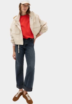 Ung kvinde iført beige jakke, rød sweater, mørke vidde jeans, brune sneakers og beige kasket står med hånden på hoften.