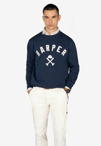 Sudadera marina con el texto blanco "HARPER" y un gráfico de hachas cruzadas. Presenta un cuello redondo y puños acanalados, combinada con pantalones de carga beige.