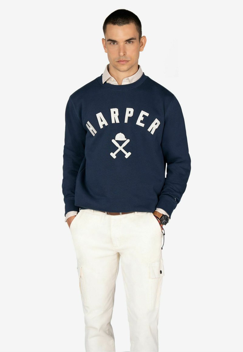 Sudadera marina con el texto blanco "HARPER" y un gráfico de hachas cruzadas. Presenta un cuello redondo y puños acanalados, combinada con pantalones de carga beige.