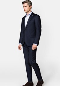 Granatowy garnitur o dopasowanym kroju, z jednolitym zapięciem na jeden guzik, klapami w kształcie notcha oraz dwiema przednimi kieszeniami. Wyraźnie biała koszula i brązowe buty.