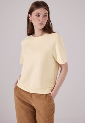 Haut jaune à manches courtes avec un col rond et des manches raglan. Texture douce, couleur unie, associé à un pantalon marron avec des accents latéraux.