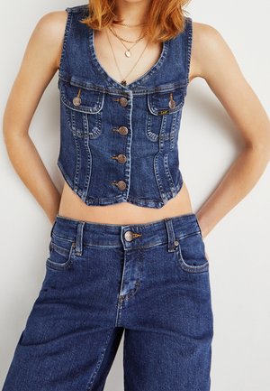 Vrouw draagt een mouwloos denimvest met knopen en high-waist denim jeans, met haar handen op haar heupen tegen een effen achtergrond.