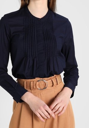 Femme portant un chemisier plissé bleu marine à manches longues, rentré dans un pantalon taille haute beige avec une large ceinture et une boucle ronde argentée.