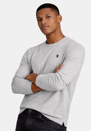 Junger Mann mit kurzen Haaren trägt ein hellgraues Langarmshirt mit kleinem schwarzem Logo, Arme verschränkt, kombiniert mit dunklen Jeans vor einfarbigem Hintergrund.