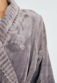 Närbild av en person som bär en mjuk, plysch lavendelfärgad badrock med en ribbad sjalkrage och en subtil broderad logotyp på bröstet.