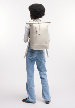 Kiharatukkainen henkilö, joka on pukeutunut valkoiseen pitkähihnaiseen yläosaan ja farkkuihin, kantaa beigeä pyörätopilla varustettua reppua, jossa on etutasku vetoketjulla.