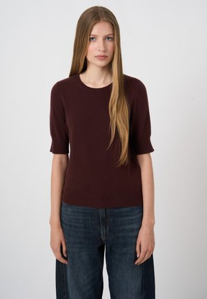 Young woman with long blonde hair wearing a dark brown short-sleeve sweater and blue jeans, standing against a plain white background.