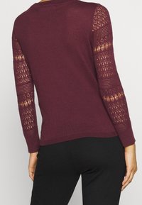 Frau trägt einen langärmligen, tief burgunderfarbenen Pullover mit gemusterten Spitzeneinsätzen an den Ärmeln und schlichtem Rücken, kombiniert mit schwarzen Hosen.
