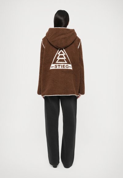 Sudadera de forro polar marrón con un gran logo de árbol blanco en la parte posterior, con capucha con cordón y ribete blanco en contraste. Llevada con pantalones negros.