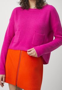 Rosa gestrickter Pullover mit gerippten Bündchen und Saum, der zwei frontale Taschen hat. Kombiniert mit einem leuchtend orangefarbenen Rock mit Reißverschluss.