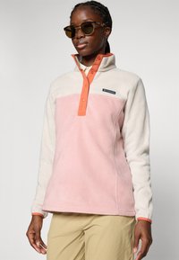 Donna che indossa un pile Columbia rosa e crema con colletto arancione a bottoni a pressione e pantaloni beige, occhiali da sole, in piedi contro uno sfondo neutro.