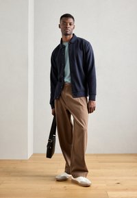 Cárdigan azul marino con cierre de botones, superpuesto sobre una camiseta verde clara, combinado con pantalones de corte relajado beige y zapatillas blancas. Sosteniendo un bolso negro.