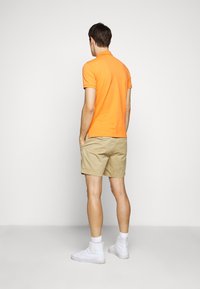 Orange pikétröja kombinerad med beige shorts, med en enkel design. Bärs med vita sneakers, sedd bakifrån.