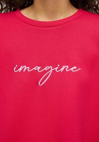 Růžové bavlněné tričko s kulatým výstřihem a krátkými rukávy, zdobené bílým kurzívním vyšíváním s nápisem "imagine" přes přední stranu.