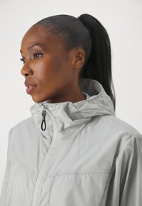 Veste coupe-vent gris clair avec un col haut, une fermeture éclair à l'avant et un tissu texturé. Comprend une boucle de suspension sur la tirette de la fermeture éclair.