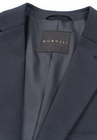 Navy blazer with a smooth texture, featuring a tailored collar and the brand label "bugatti" in gold on a dark background.