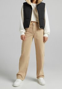 Black puffy vest with beige lining, white cropped sweatshirt, and beige wide-leg pants. White sneakers complete the outfit.