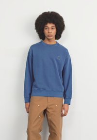 Sudadera azul de algodón, con cuello redondo, puños acanalados y un pequeño parche con logo en el pecho. Combinada con pantalones beige.