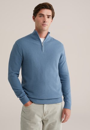Suéter de color azul claro con cuello con cremallera, patrón de punto texturizado y puños y dobladillo acanalados. Llevado con pantalones de color beige claro.