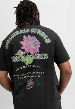 Black cotton t-shirt featuring a colorful floral graphic, text in bold green and white, and various accents including a logo and year.