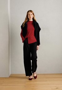 Red long-sleeve top with a knotted accent, paired with black wide-leg trousers. Model wears red flat shoes. Neutral background and wooden floor.