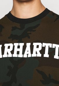 Olijfgroene camouflage T-shirt met korte mouwen en een zwarte geribbelde halslijn, met gedurfde, witte "CARHARTT" tekst over de borst.
