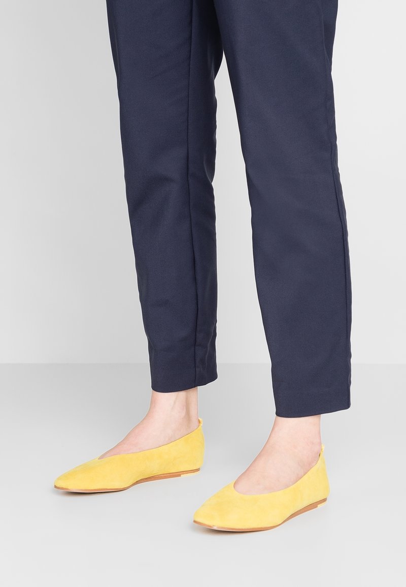 Chaussures plates en daim jaune avec un design à bout pointu, associées à un pantalon tapered bleu marine. Les chaussures présentent une silhouette minimaliste et un profil bas.