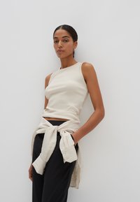 Débardeur côtelé beige avec un col montant, associé à un pantalon noir décontracté et une chemise claire nouée autour de la taille.