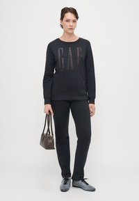 Sudadera negra con el logo "GAP" en el frente, combinada con pantalones negros. Zapatillas grises y un bolso pequeño de cuero marrón oscuro completan el atuendo.