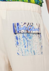 Bézs nadrág, melynek zsebe absztrakt kék-fehér mintázattal texturált. A zseben egy kék címke található, melyen a "Carlo Colucci" felirat olvasható.