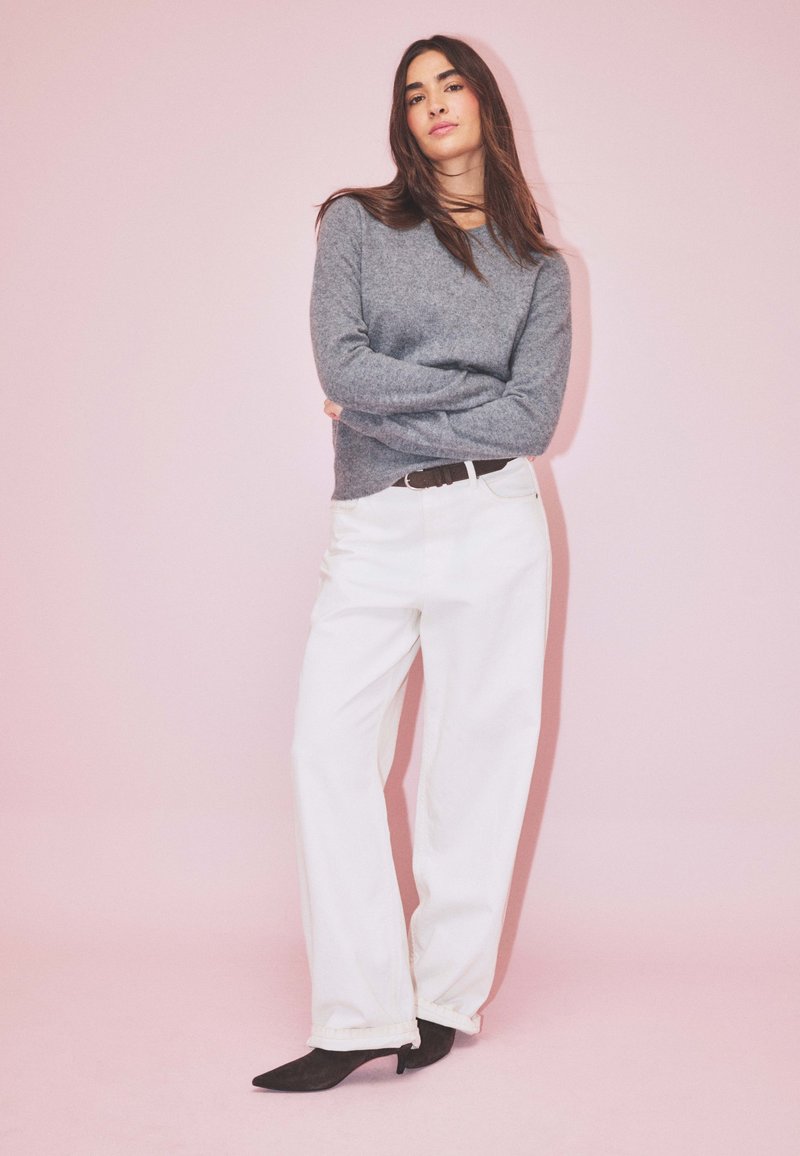 Un pull en laine gris avec un col rond, associé à un pantalon large blanc et des escarpins noirs à bout pointu, devant un fond rose.
