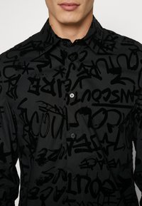 Camicia nera con bottoni, caratterizzata da un motivo graffiti nero lucido, composto da varie forme astratte e lettere, realizzata in un tessuto morbido.