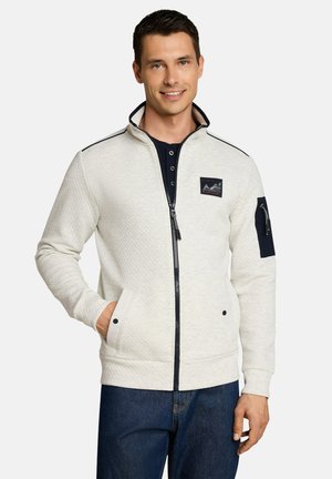 Smiling man wearing a light quilted zip jacket with dark trim and patch on chest and sleeve, standing with one hand in pocket.