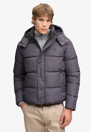 Junger Mann trägt eine dunkelgraue Steppjacke über einem grauen Reißverschluss-Pullover und beigen Hosen und steht mit einer Hand in der Tasche.