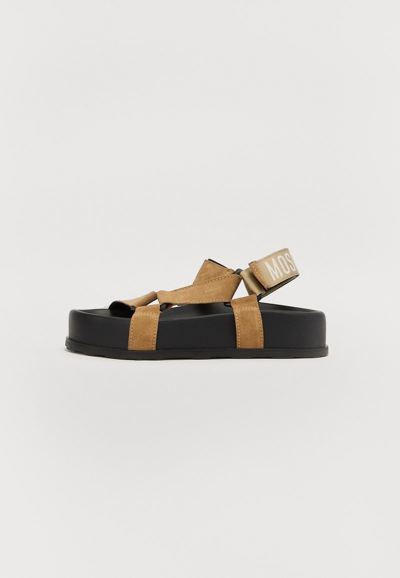 Brown suede sandals with a thick black sole, featuring cross straps and a branded strap on the heel. Flat design with closed toe.