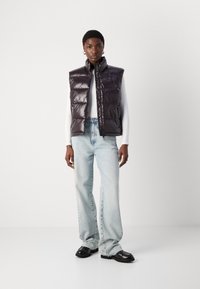 Prsluk od puha u tamnoljubičastoj boji, s visokim ovratnikom i prednjim džepovima. Uparen s svijetloplavim trapericama ravnog kroja i crnim cipelama.