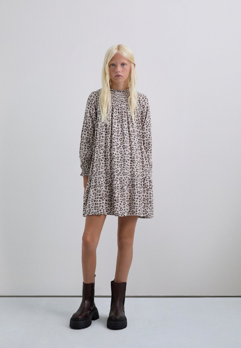 Vestido recto beige con estampado floral, de mangas largas y escote fruncido, combinado con botas negras de tobillo grueso. Textura suave por toda la prenda.