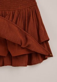 Rostfarbener, geschichteter Rock mit dezenten goldenen vertikalen Streifen und elastischem Bund, der innere Shorts unter der gerafften Außenschicht zeigt.